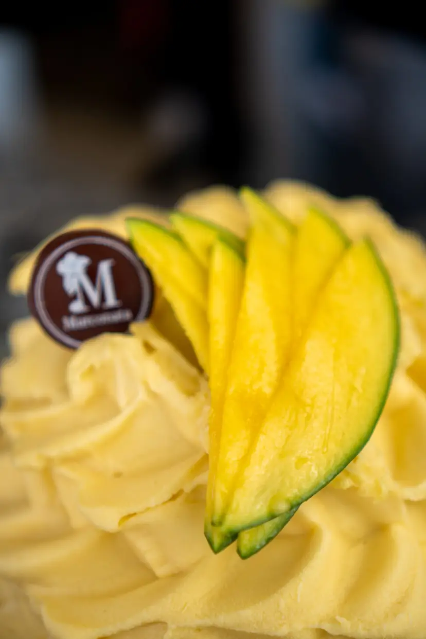 Helado amarillo con láminas de mango por encima y una placa de chocolate con la M de Marconata