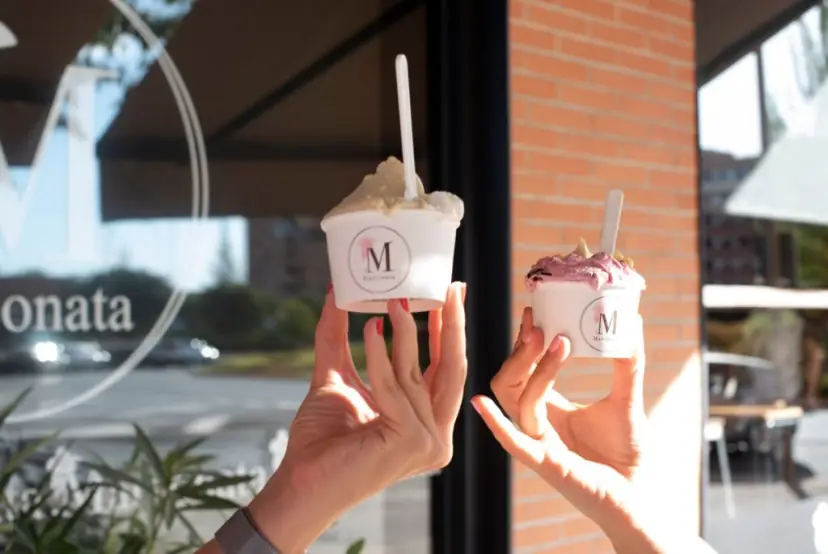 Dos manos levantan tarrinas de Marconata, una con helado claro y otra con helado morado, ante la cristalera rotulada del local