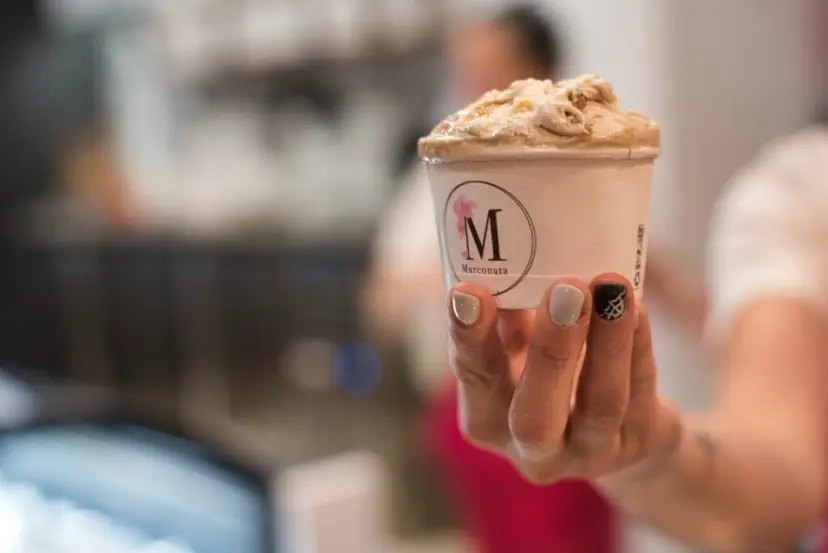Una empleada con delantal rojo tiende hacia ti una tarrina de Marconata colmada de helado de color café