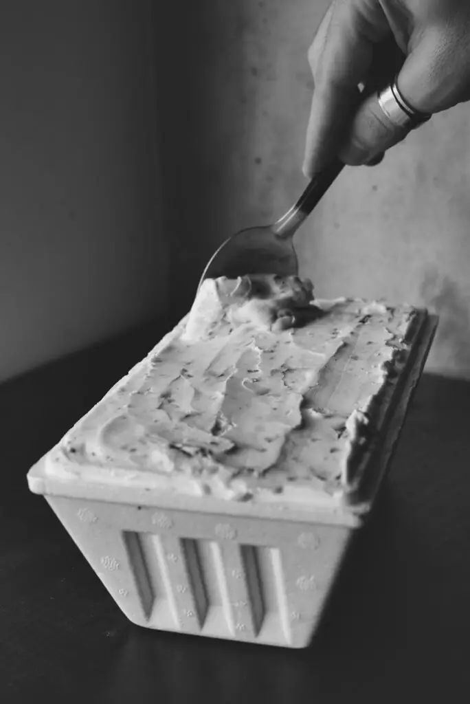 Una mano hunde una cuchara en una barqueta de helado para llevar, sobre una mesa oscura